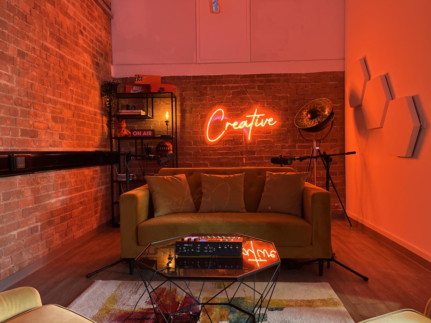 A podcast studio with exposed brick walls, neon 'Creative' sign, mustard sofa, and recording equipment at Creators House.