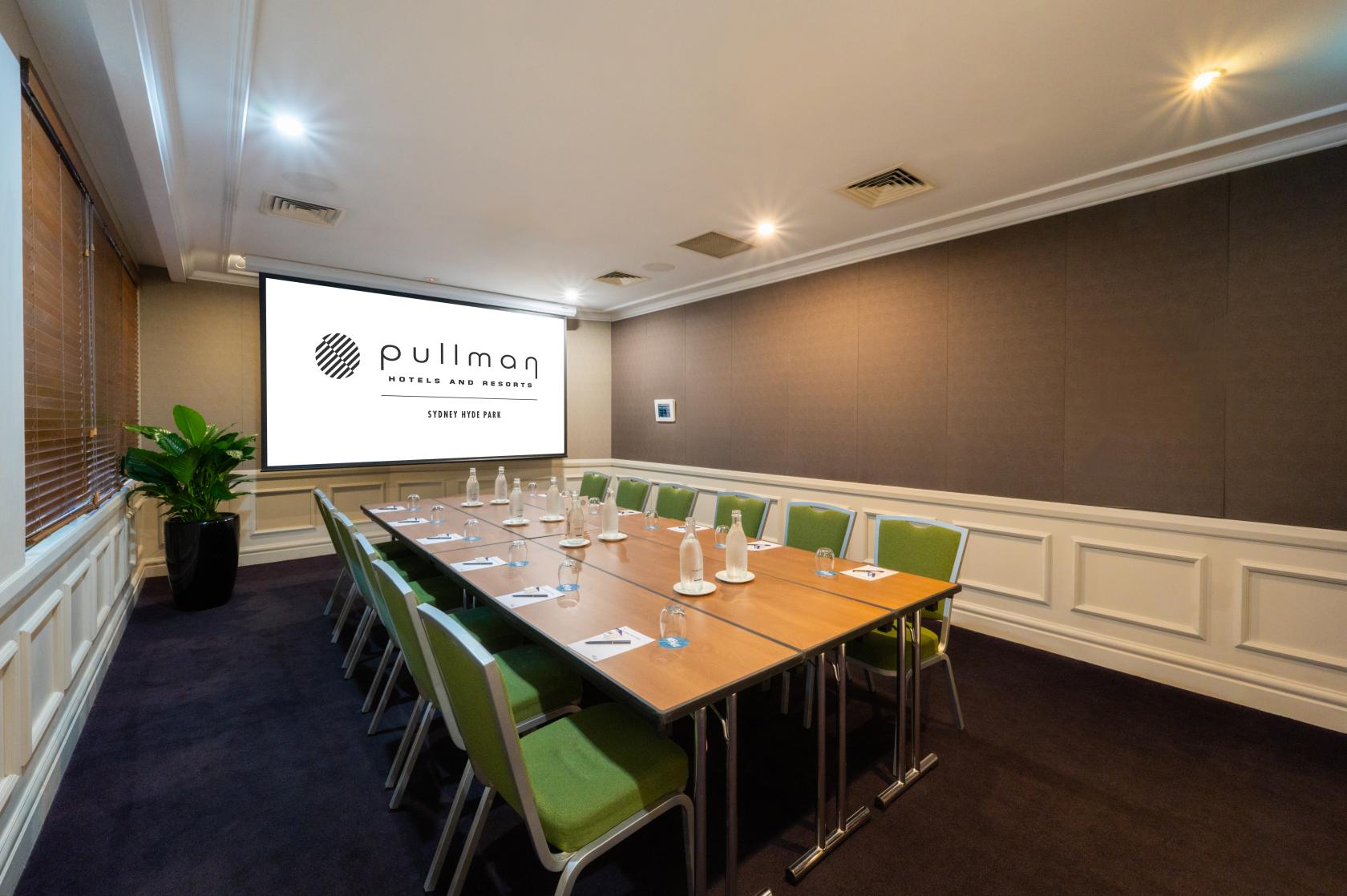A meeting room with a long conference table, green chairs, and a large presentation screen at Pullman Sydney Hyde Park.