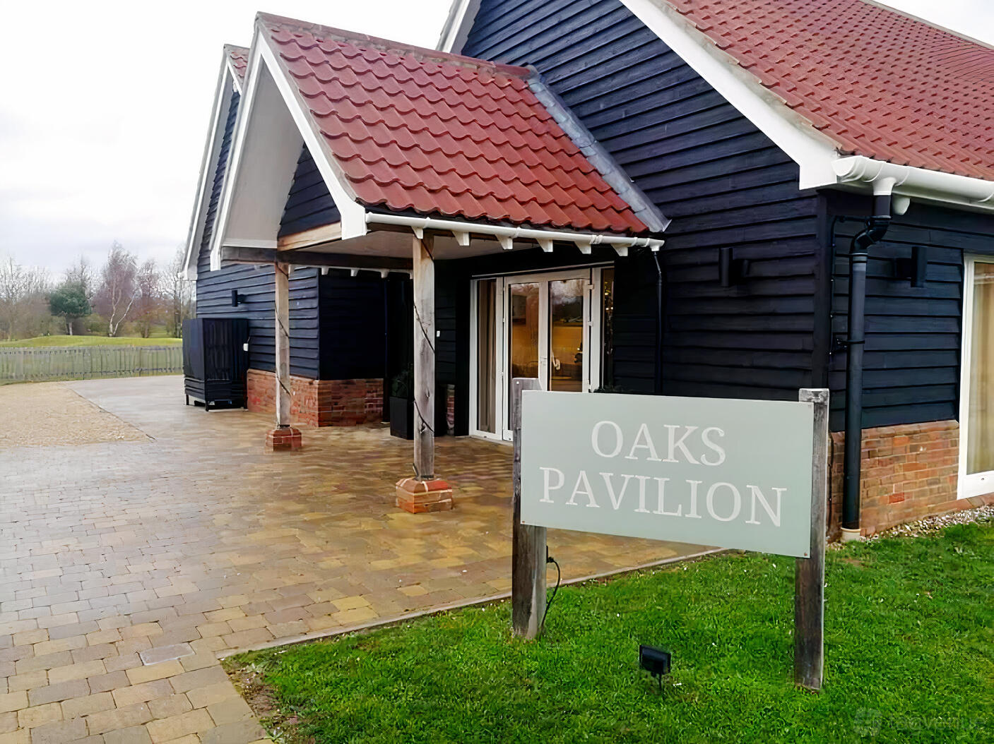 A golf club exterior with dark wood siding, red roof tiles, and a sign for Oaks Pavilion at Rivenhall Oaks Golf Centre.