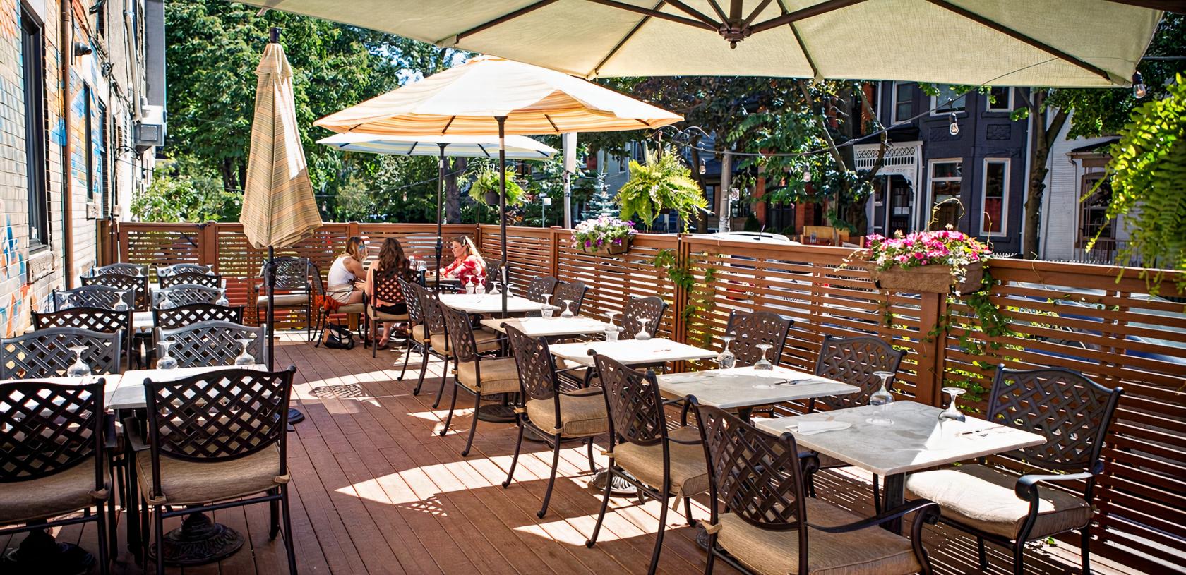 An outdoor patio with umbrella-shaded tables, potted flowers, and wood privacy fencing at Her Father's Cider Bar + Kitchen.
