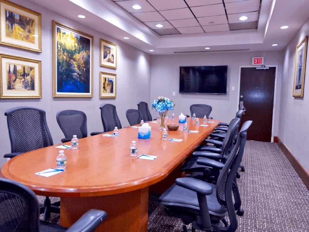 A meeting room with a long wooden conference table, black mesh chairs, wall art, and a wall-mounted TV at Harmony Suites Secaucus Meadowlands.