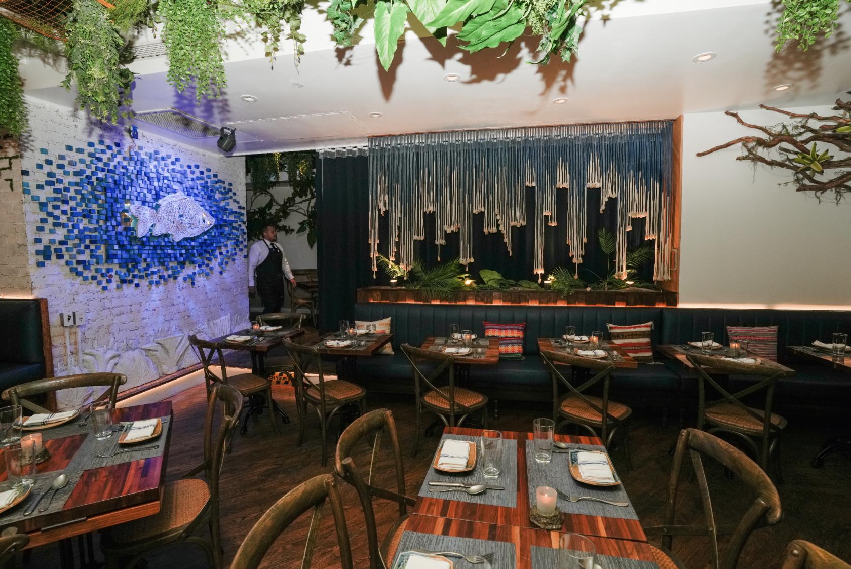A restaurant dining room with mosaic fish wall art, hanging greenery, and wooden tables at Mission Ceviche.