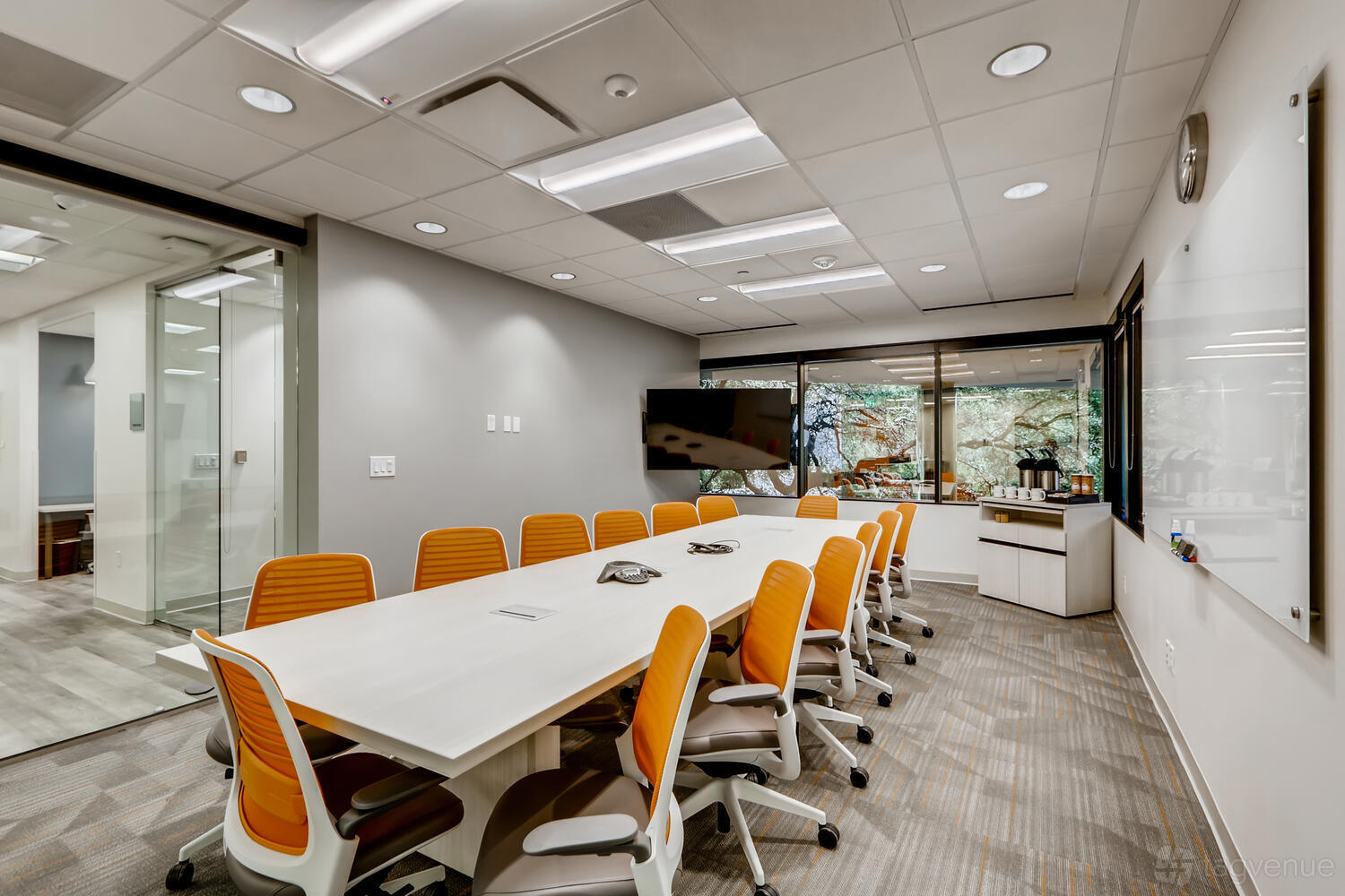 A boardroom with a long conference table, orange chairs, wall-mounted TV, and windows at Office Evolution - Austin Arboretum.