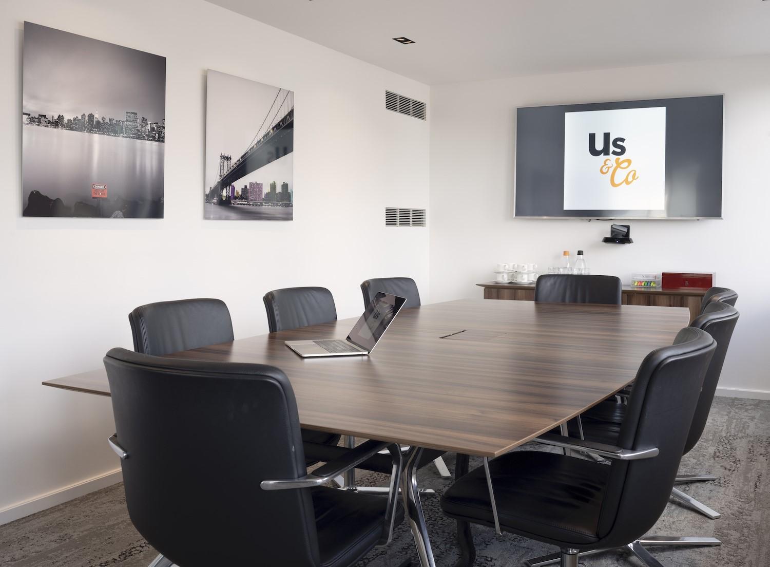 A boardroom with a wooden conference table, black leather chairs, wall art, and a mounted screen at Us&Co Monument.