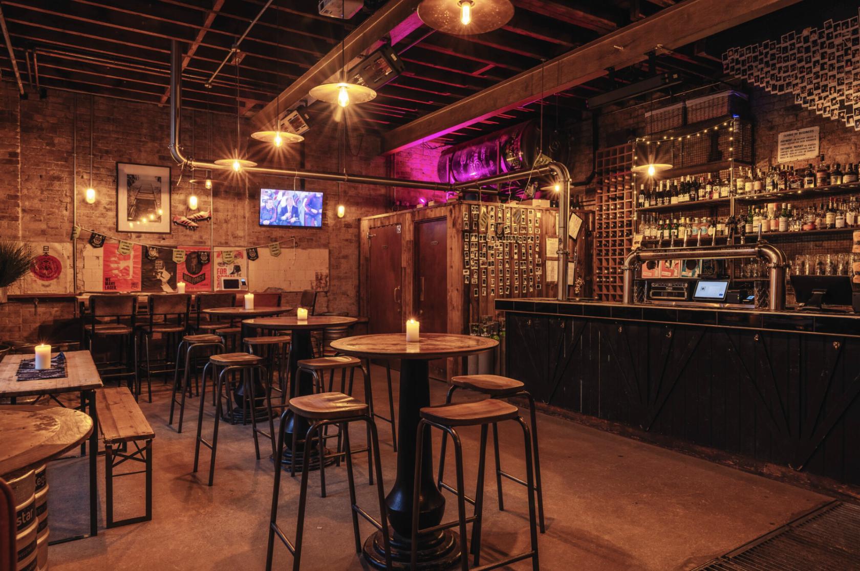 A pub with exposed brick walls, industrial lighting, wooden tables, and a stocked bar at Wolfpack Queens Park.