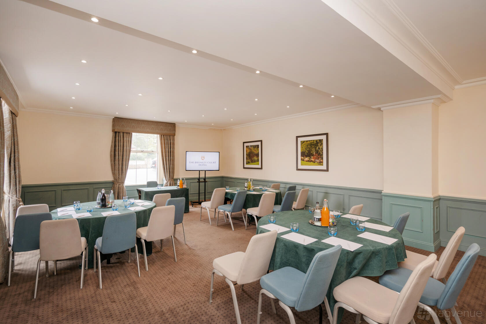 A meeting room with natural light, green tablecloths, a presentation screen, and framed artwork at The Bromley Court Hotel.