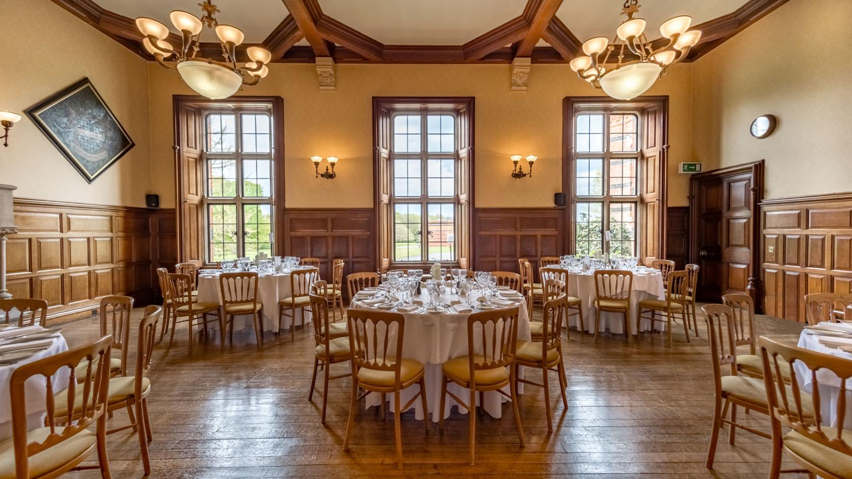 A hotel event room with wood-paneled walls, large windows, and round tables set for dining at The Elvetham Hotel.