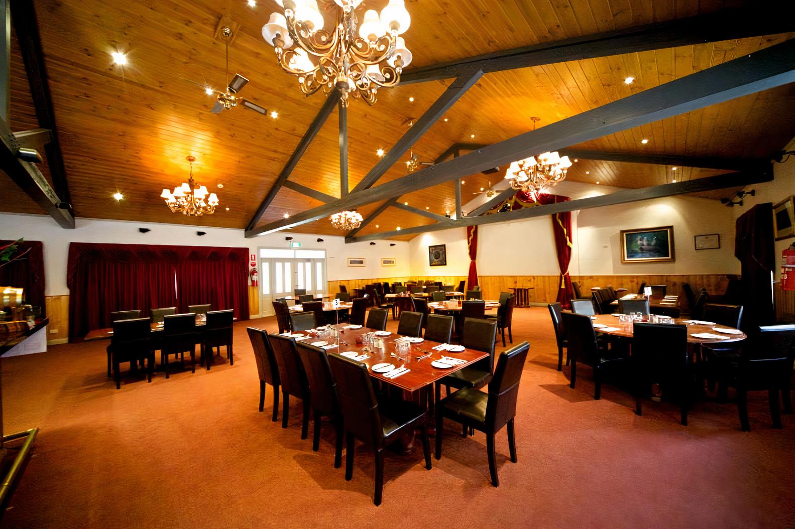 A restaurant with wooden beam ceilings, chandeliers, large windows with red curtains, and arranged dining tables at Yarra Valley Restaurant.