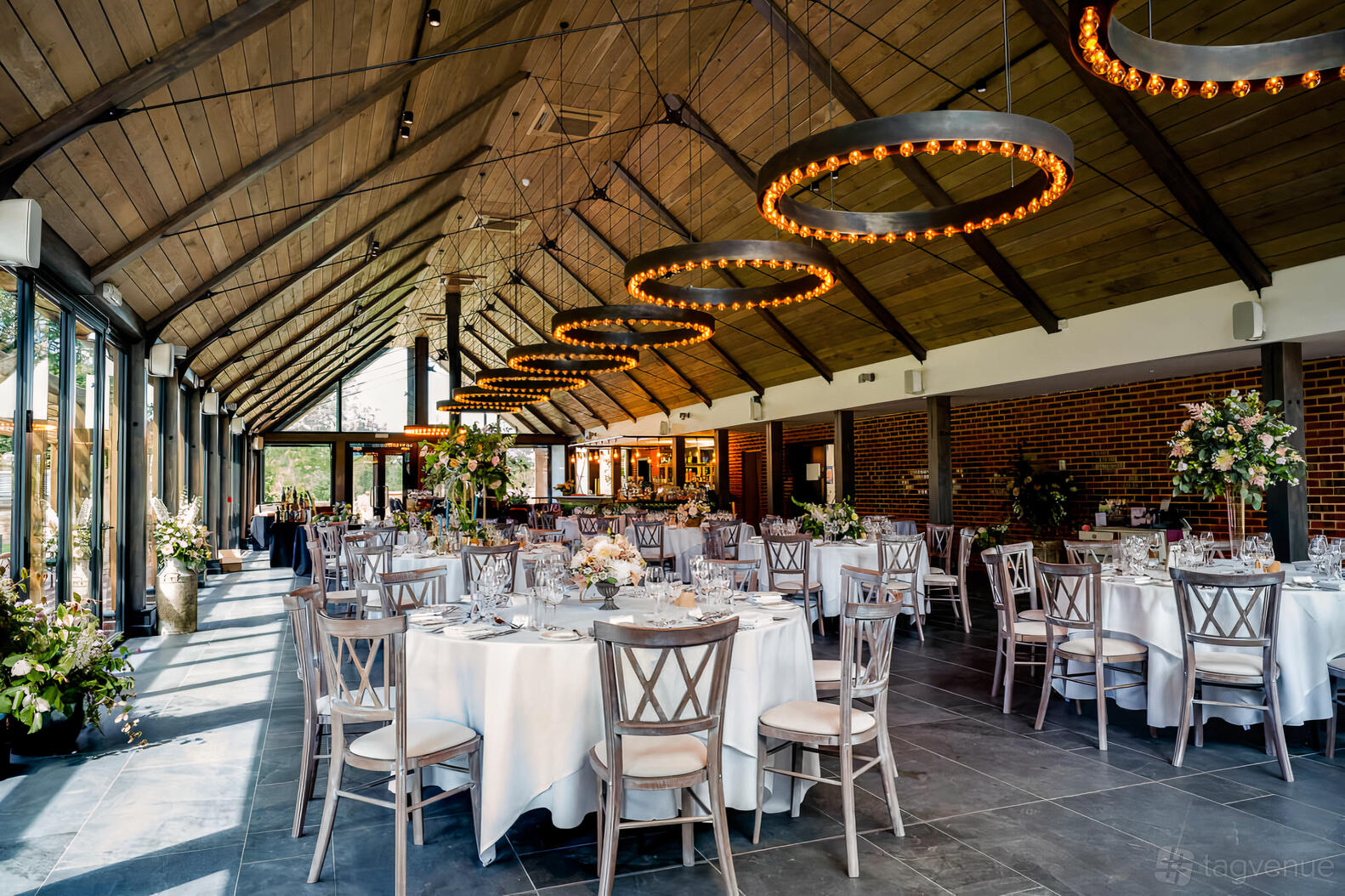 An event venue with vaulted wood ceiling, exposed brick walls, and round tables with white linens at Syrencot.