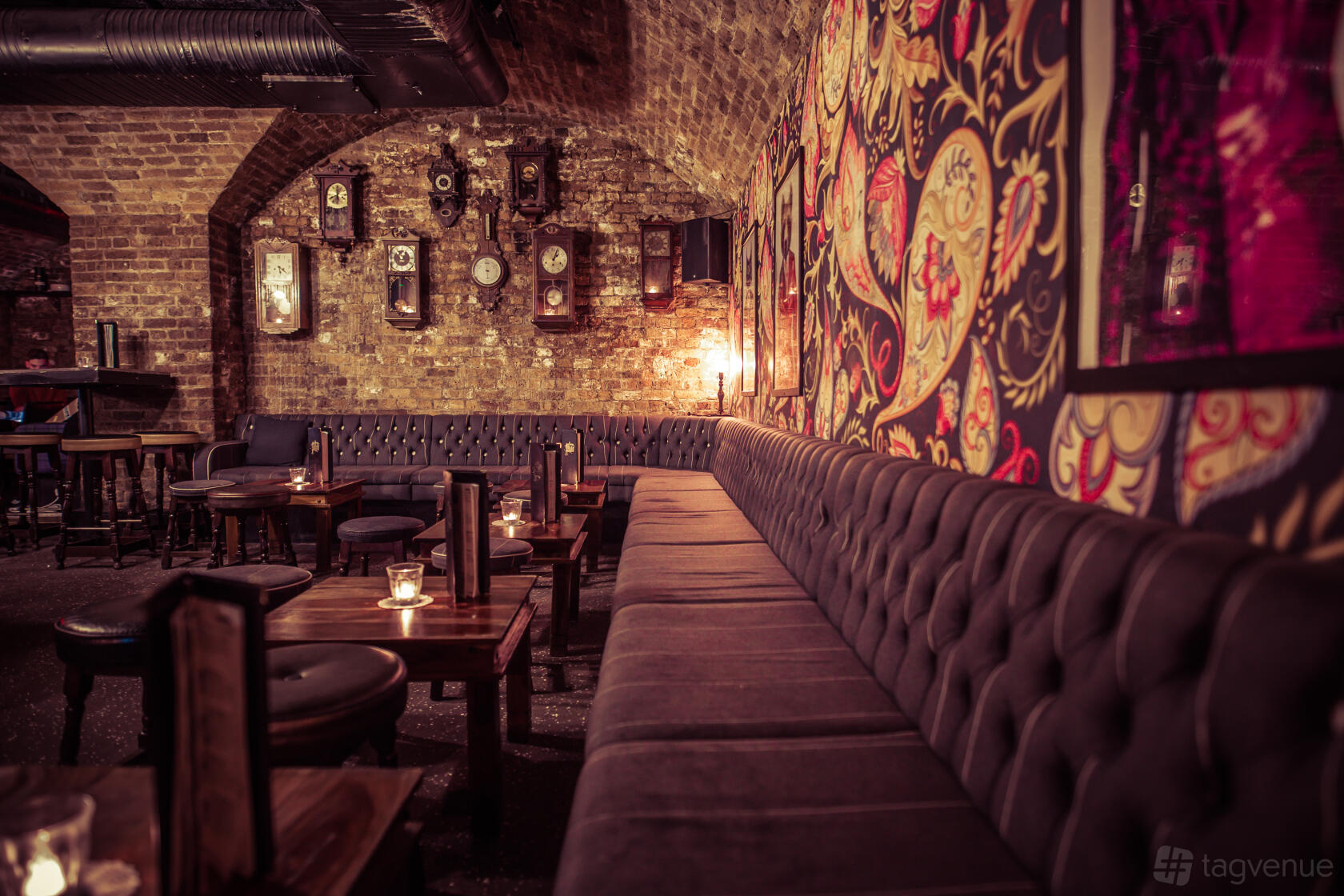 A cocktail bar with exposed brick walls, patterned wallpaper, vintage clocks, and tufted banquette seating at The Cocktail Club Monument.