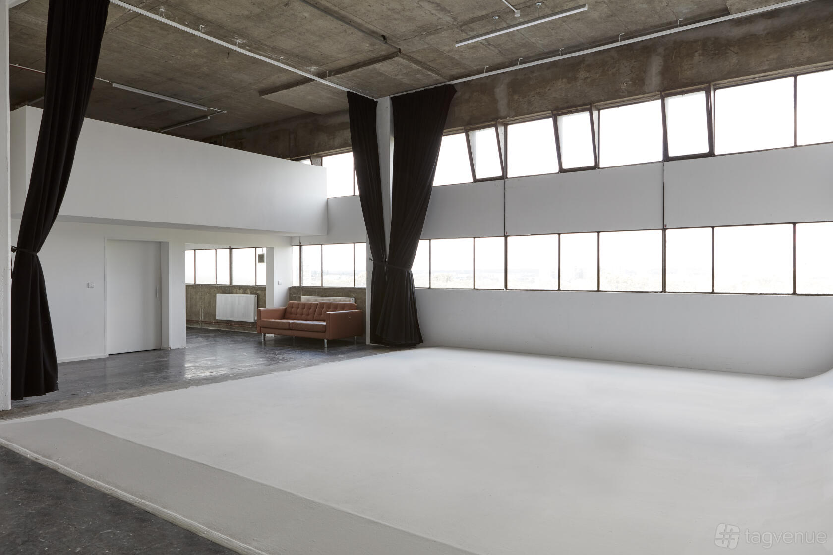 A photo studio with a large cyclorama wall, concrete ceiling, wall of windows, and brown sofa at Studio Monde.