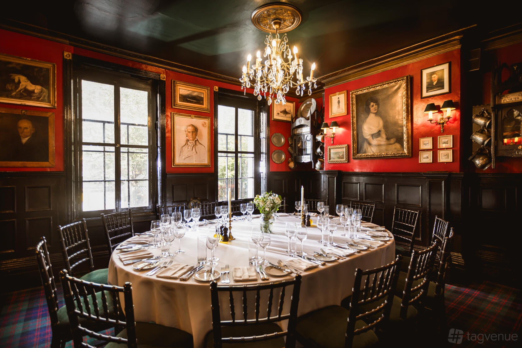 A dining room with a round table set for a meal, red walls, chandeliers, and framed portraits at Boisdale of Belgravia.