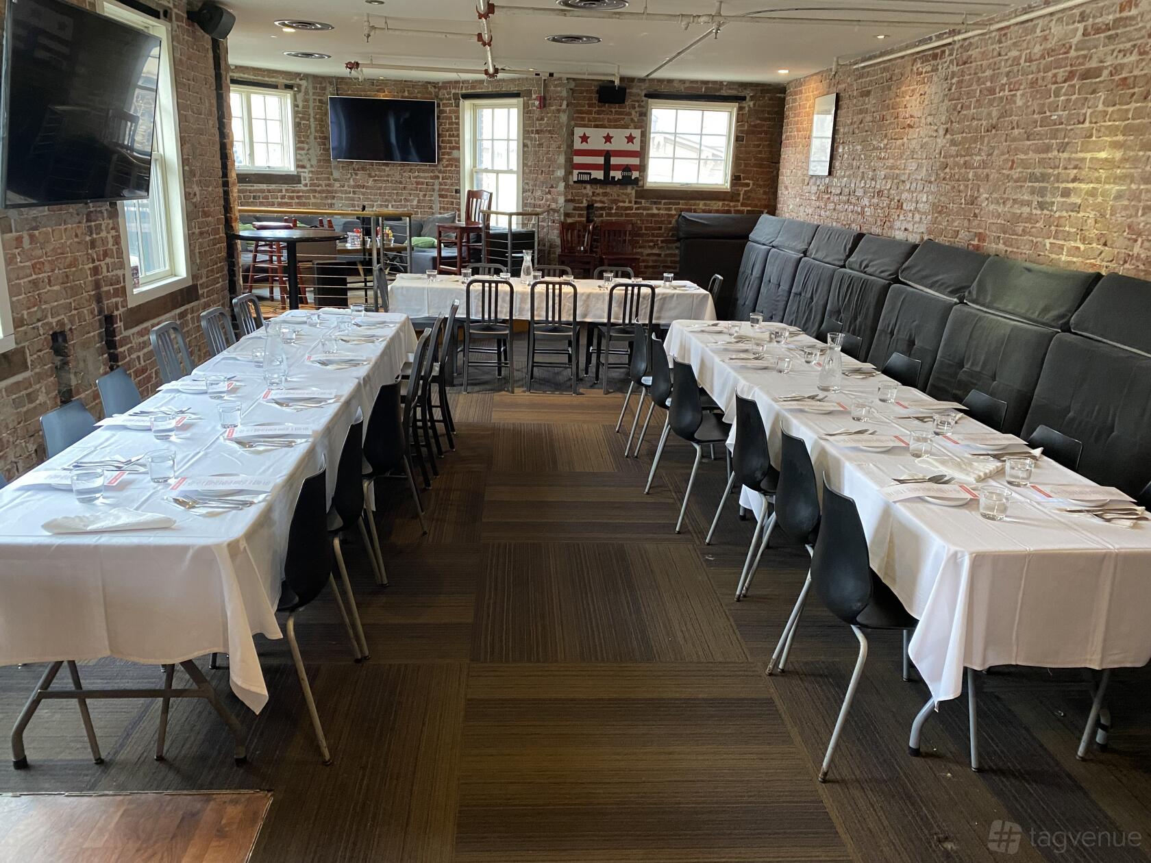 A lounge with exposed brick walls, arranged rectangular tables with white tablecloths, and black chairs at Tonic At Quigley’s Bar and Restaurant.