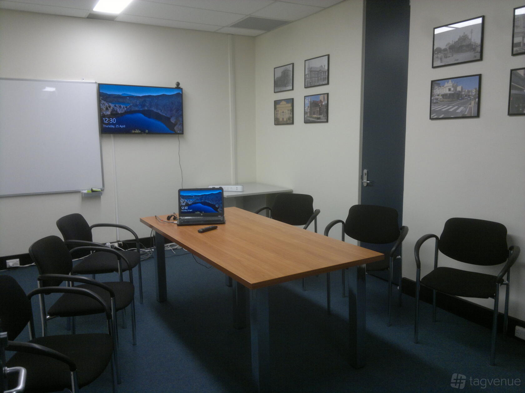 A boardroom with a rectangular wooden table, black chairs, a wall-mounted screen, and framed photos at Hot Desk Heaven.