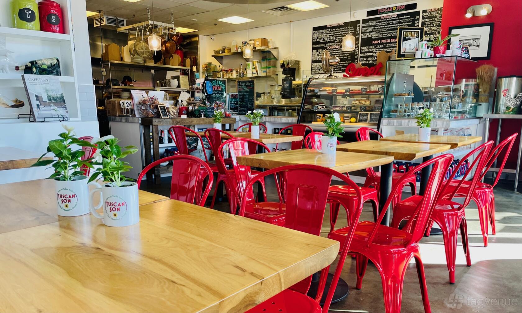 A restaurant with wooden tables, red metal chairs, potted plants, and a chalkboard menu at Tuscan Son.