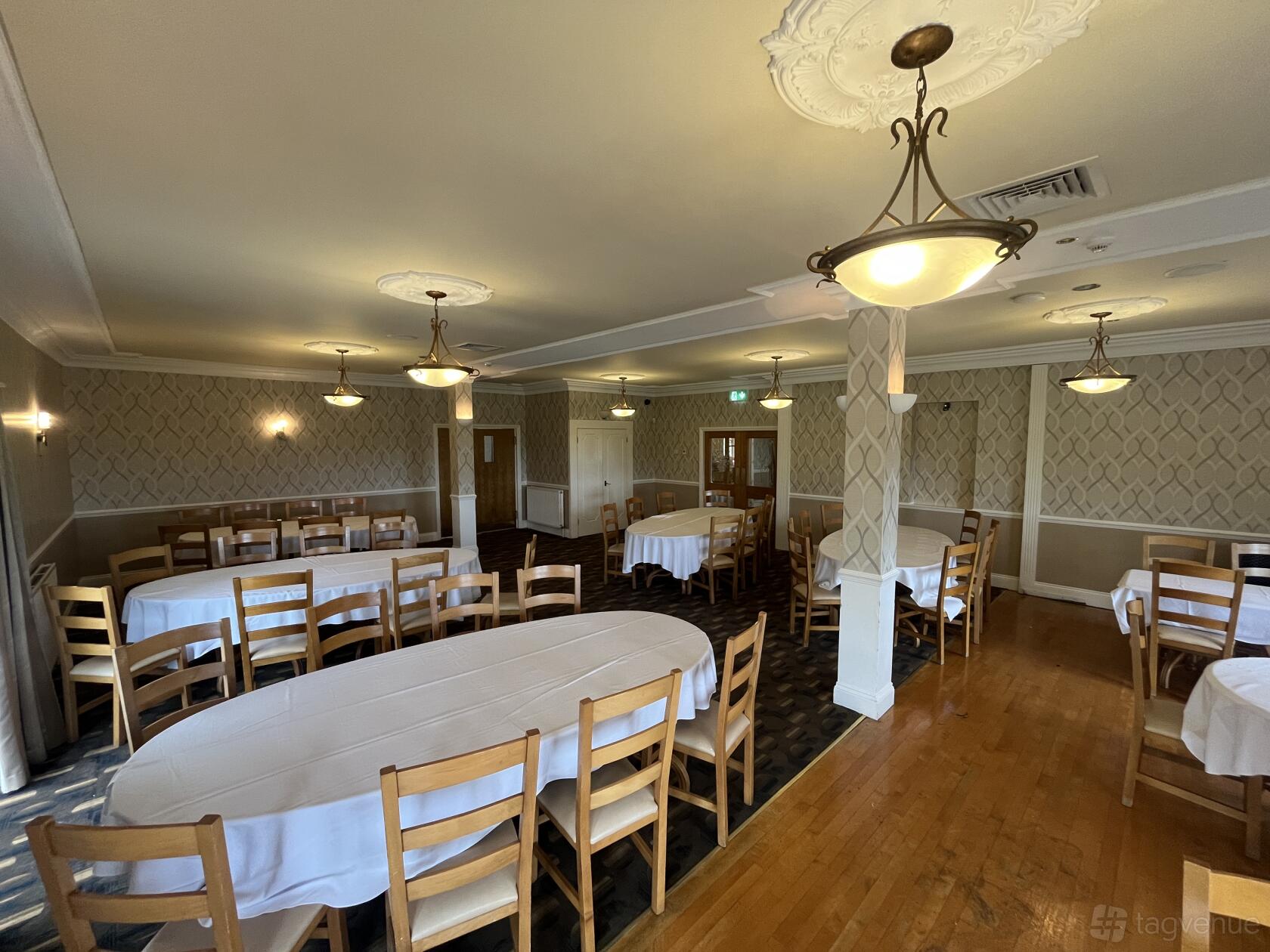 A function room with wooden chairs, oval and rectangular tables with white linens, and patterned wallpaper at The Manor Golf Club.