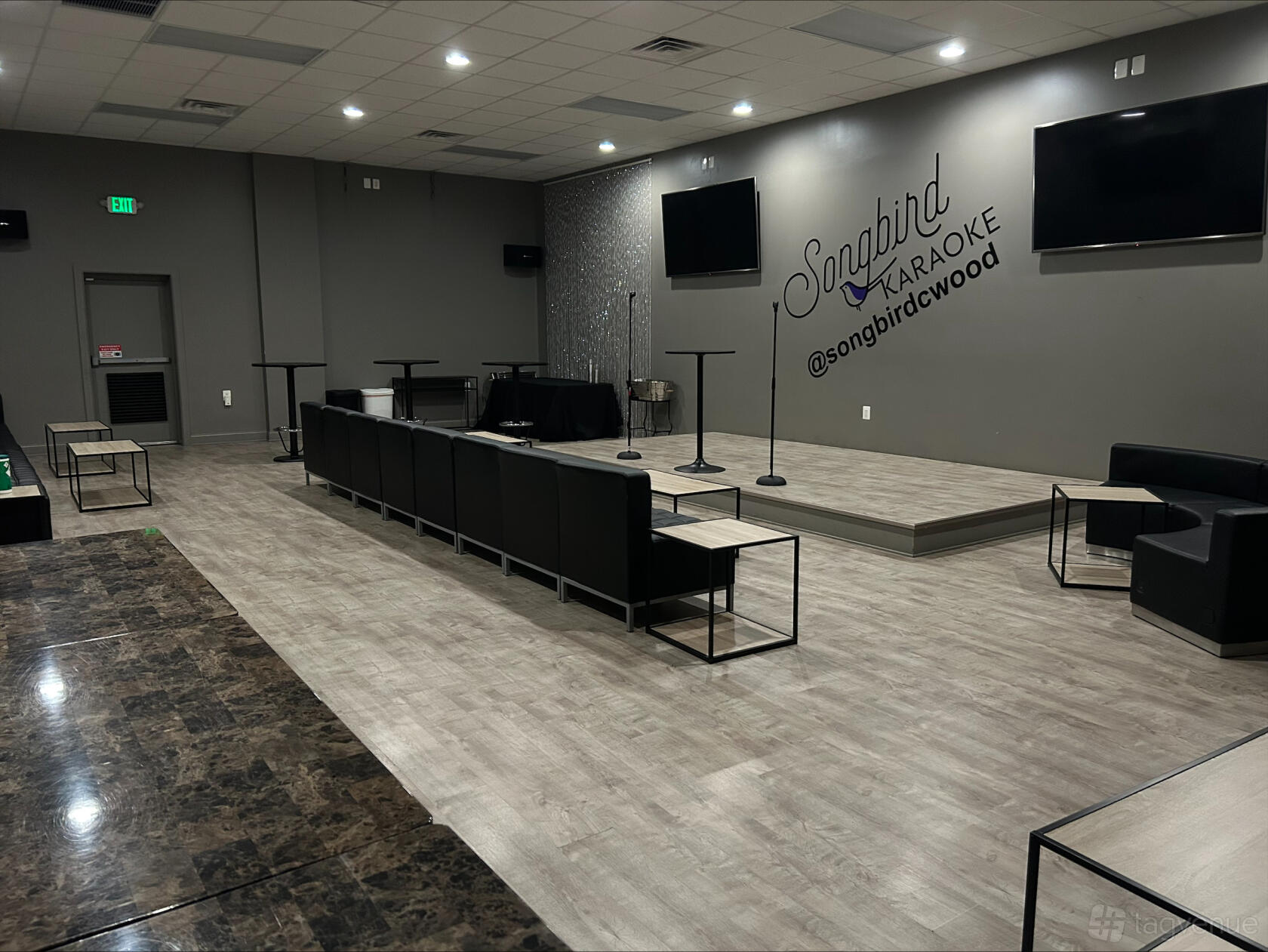 An event space in a karaoke bar with a small stage, microphone stands, lounge seating, and wood floors at Songbird Karaoke.