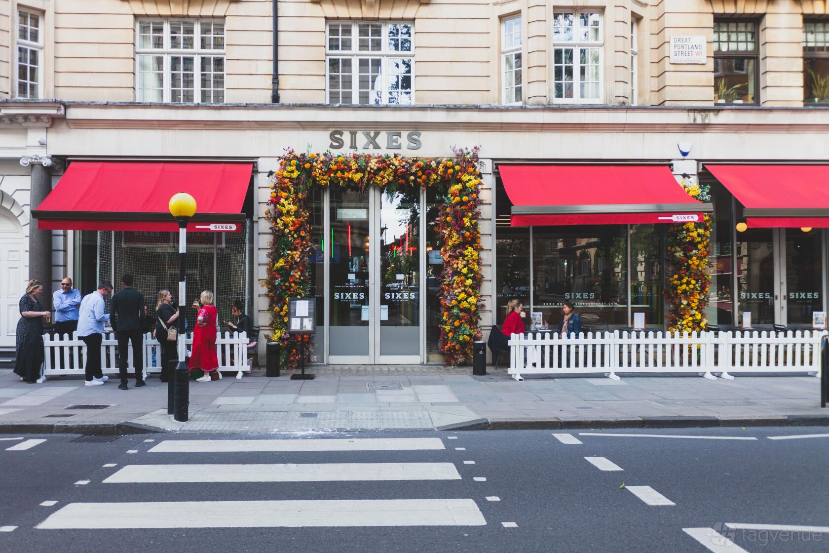 A versatile event space with large windows, red awnings, and a flower-adorned entrance at Sixes Social Cricket - Fitzrovia.