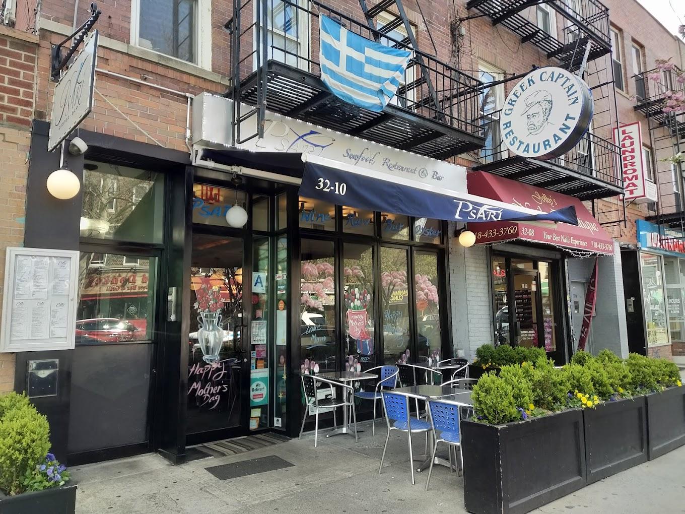 A restaurant with an outdoor sidewalk seating area, awning, and Greek flag at Psari Restaurant.