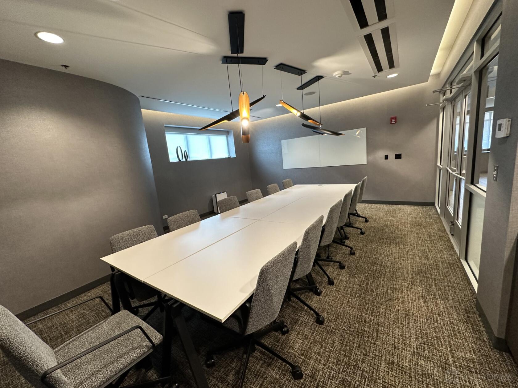 A hotel boardroom with a long white conference table, gray chairs, a glass wall, and a wall-mounted whiteboard at SpringHill Suites Chicago Chinatown.