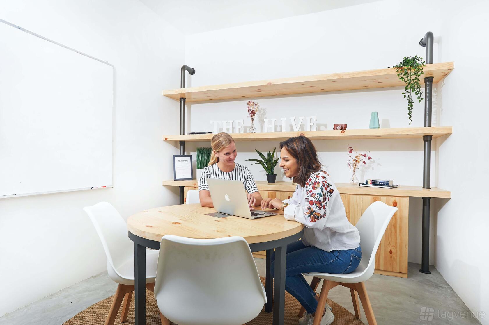 A meeting room with a round wooden table, four white chairs, wall shelves, and a whiteboard at The Hive Lavender.