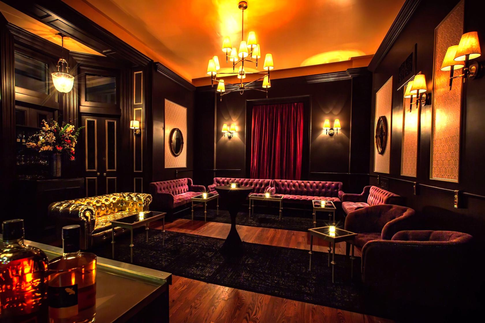 A cocktail bar with velvet tufted sofas, gold accents, and dim chandelier lighting at The Raines Law Room at The William.