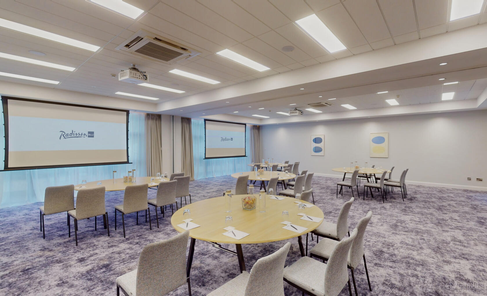A meeting room with round tables, grey chairs, projection screens, and soft carpet at Radisson Blu Hotel, Dublin Airport.