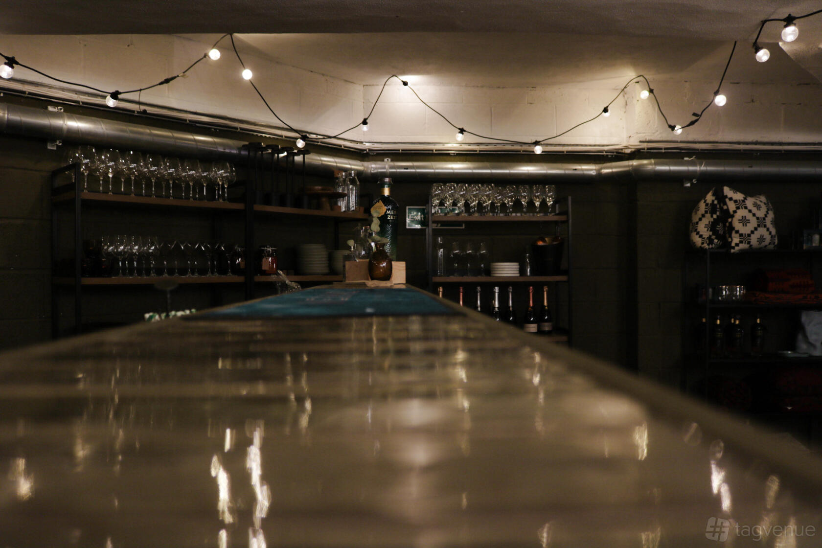 A basement event venue with exposed pipes, shelves of glassware, and string lights at Brityard.