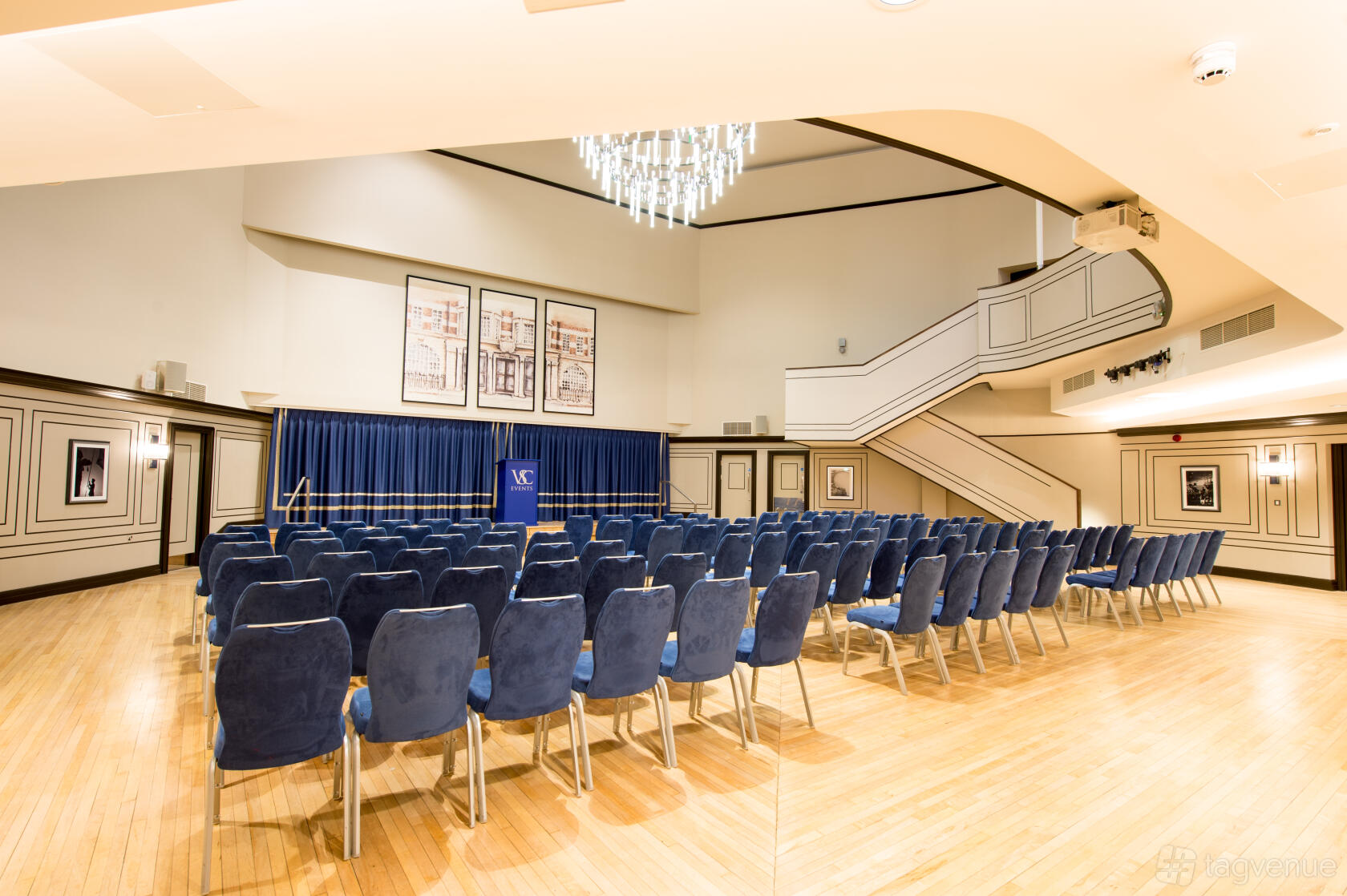 A spacious hall with tiered seating, blue chairs, wood flooring, large chandelier, and framed artwork at Victory Services Club.