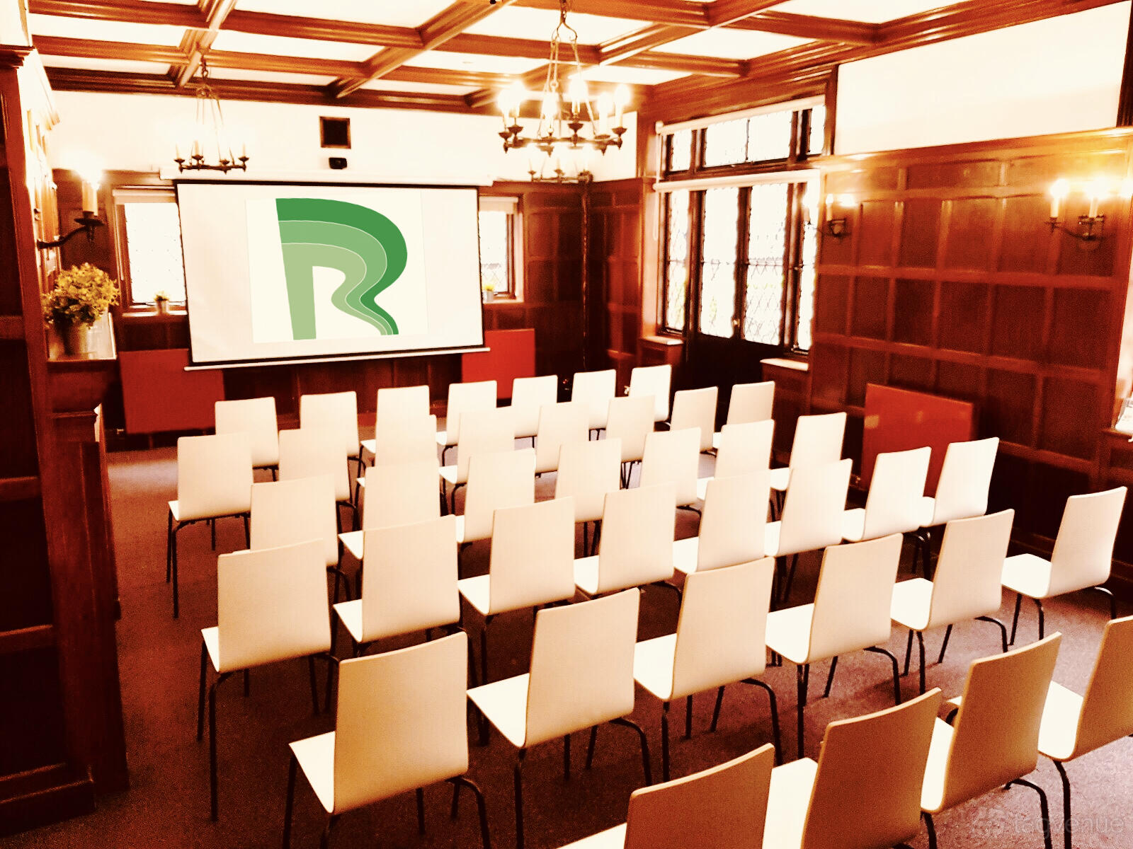 A meeting room with rows of white chairs, chandeliers, wood-paneled walls, and a projector screen at Rivertech.