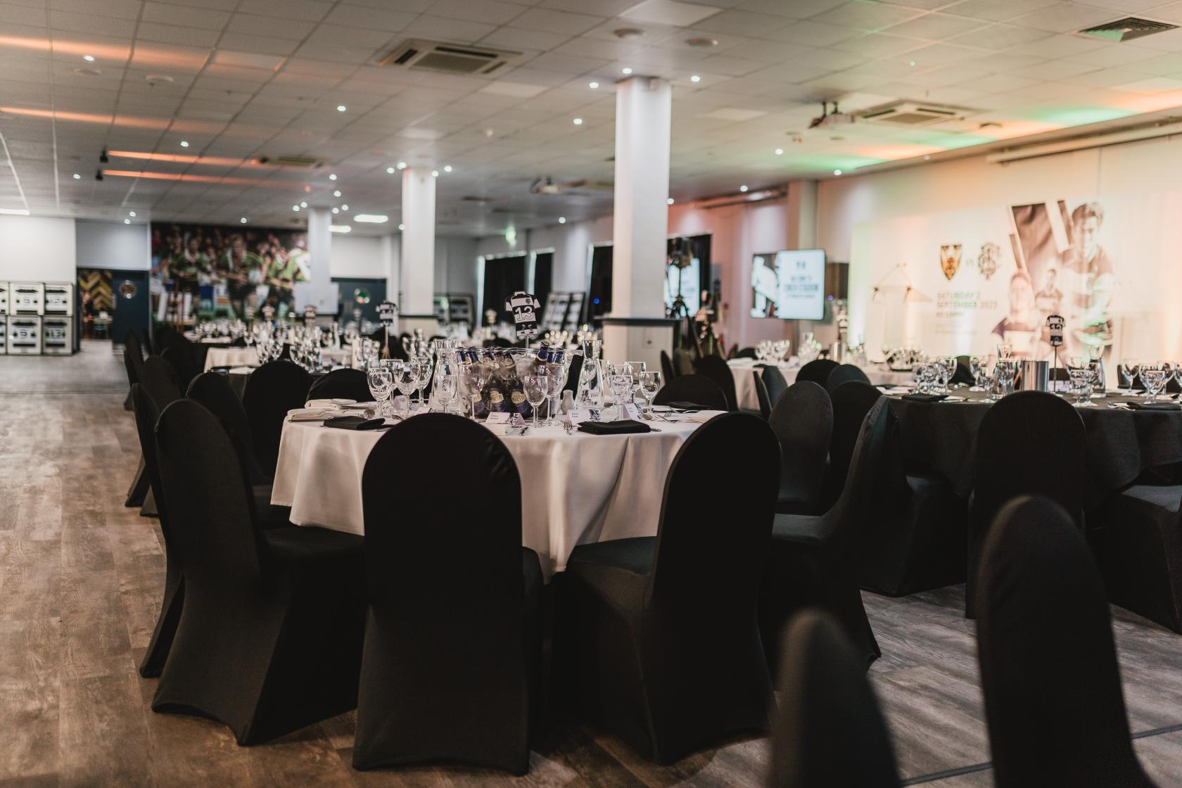 An event space with round tables covered in white linens and black chairs at Northampton Saints Stadium.