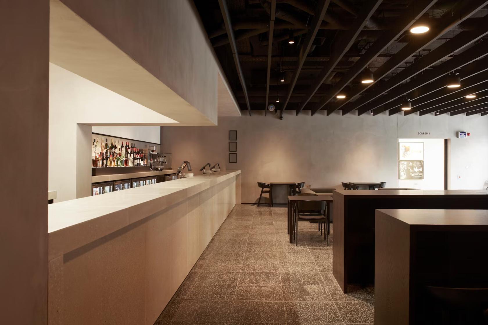 A cinema bar with a long stone counter, backlit shelves of bottles, and minimalist black tables at Curzon Hoxton.