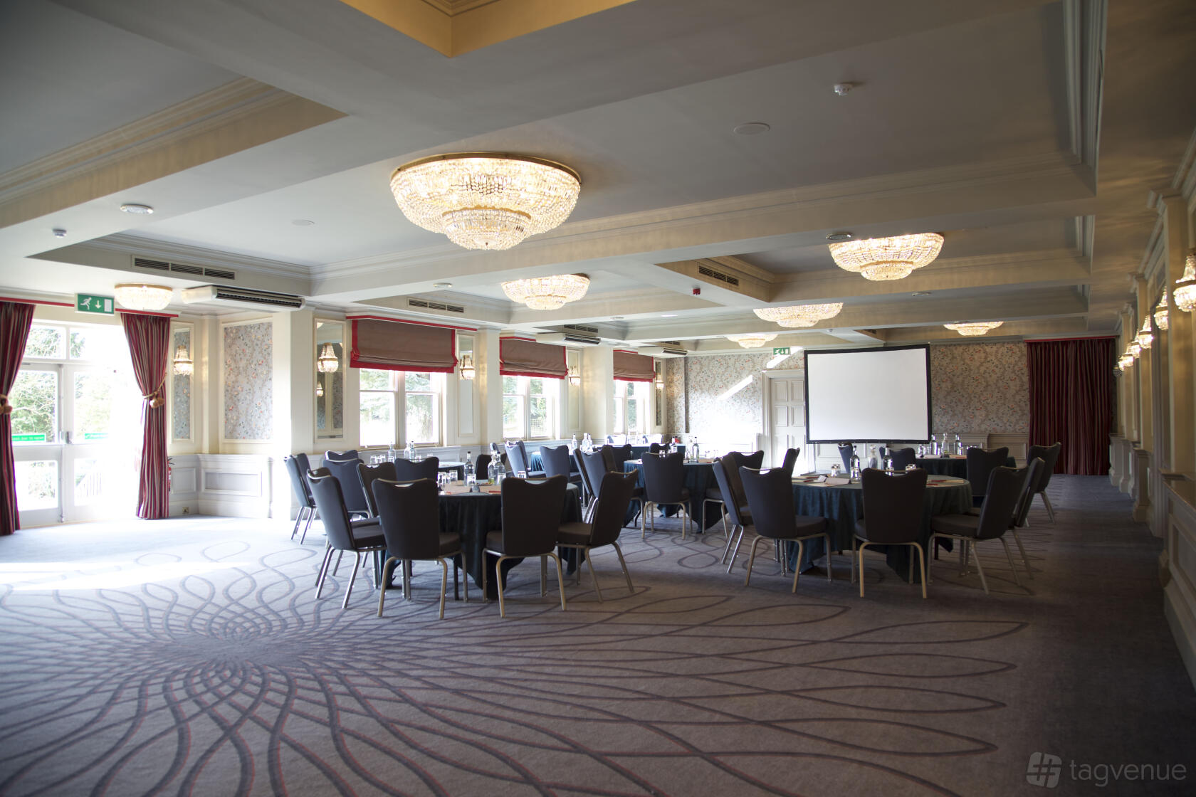 A ballroom with round tables, upholstered chairs, chandeliers, carpeted floors, and a projector screen at Pendley Manor Hotel.