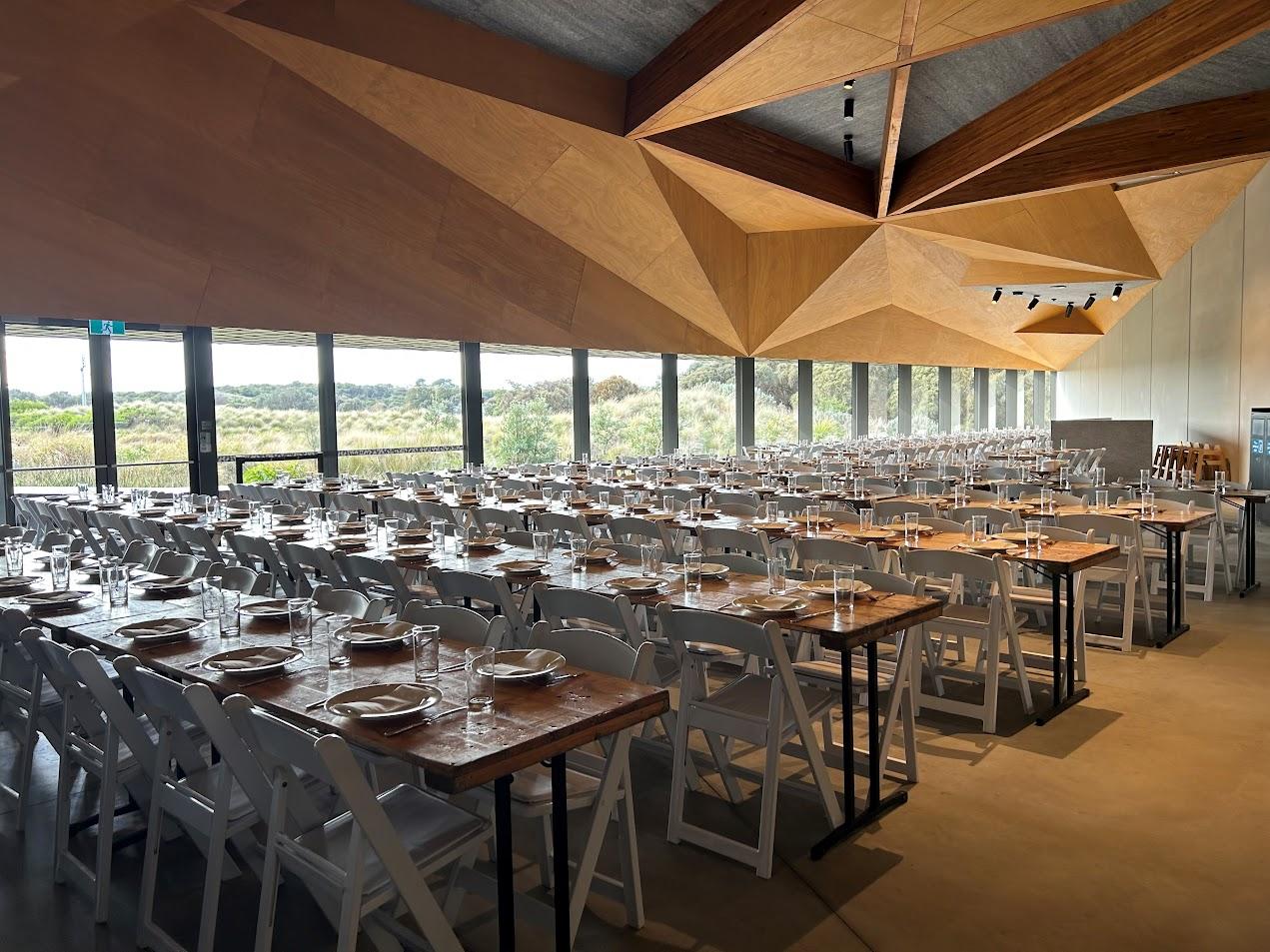 An event venue with floor-to-ceiling windows, wooden geometric ceiling, and rows of set tables at Penguin Parade Centre.