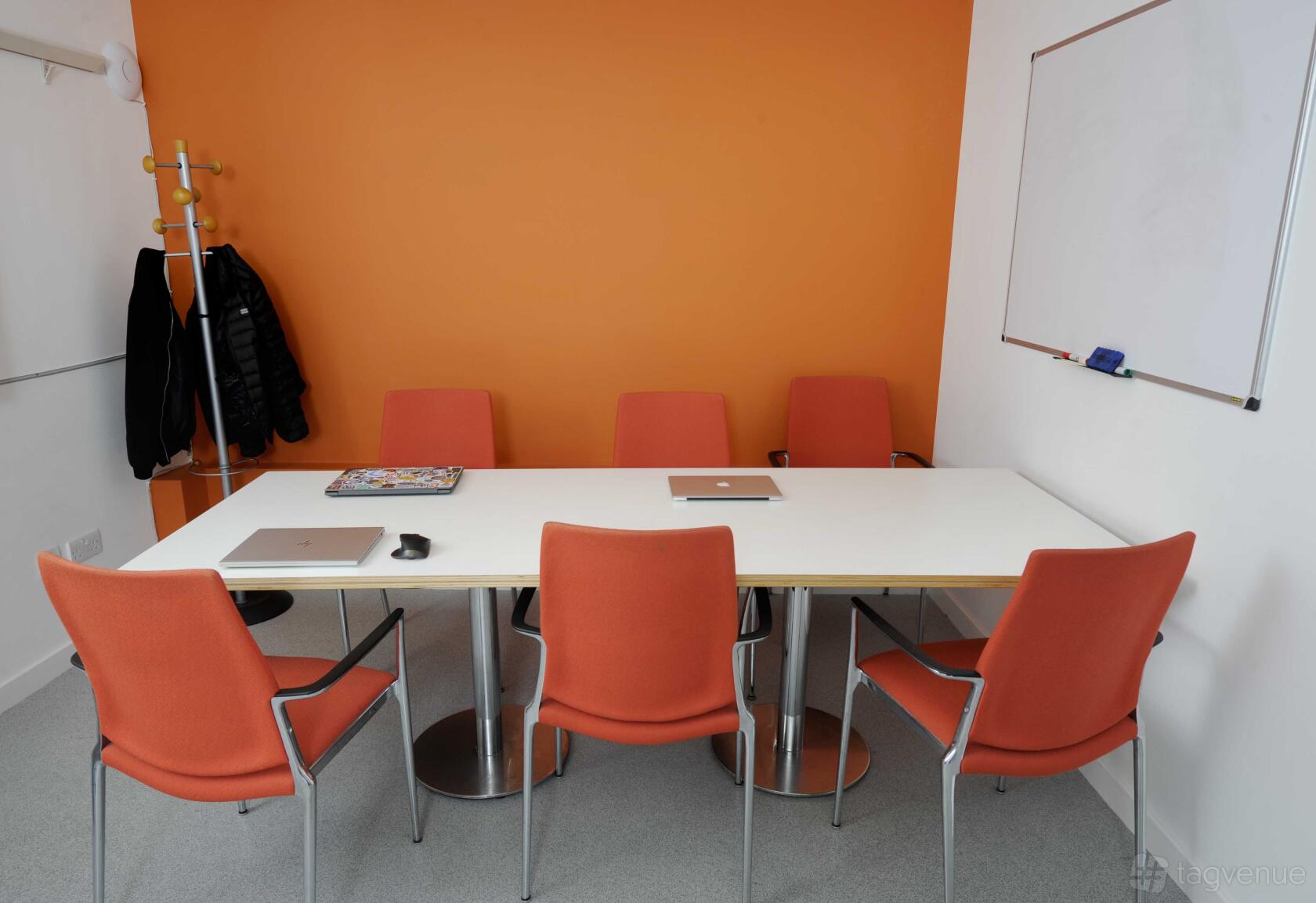 A meeting room with orange chairs, a white table, a whiteboard, and an orange accent wall at The Melting Pot.