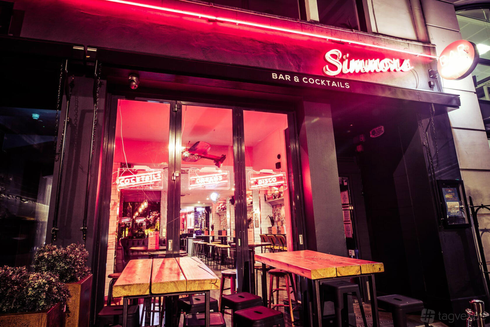 A bar with neon lighting, outdoor wooden tables, and indoor bar seating at Simmons | Piccadilly Circus.
