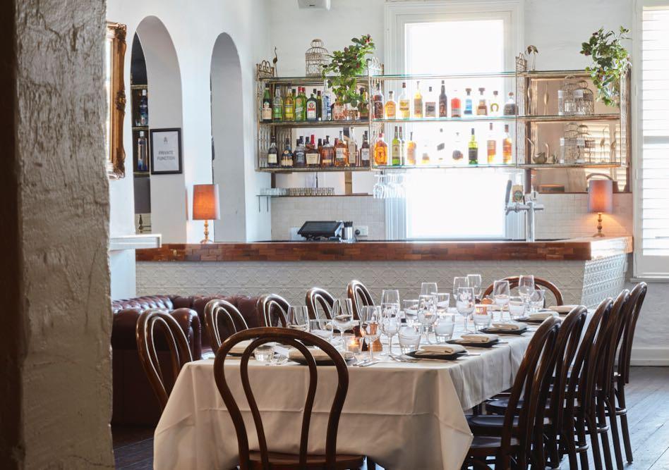 A function room in a pub with a stocked backlit bar, arched windows, and a long table set with white linens at Aviary Hotel.