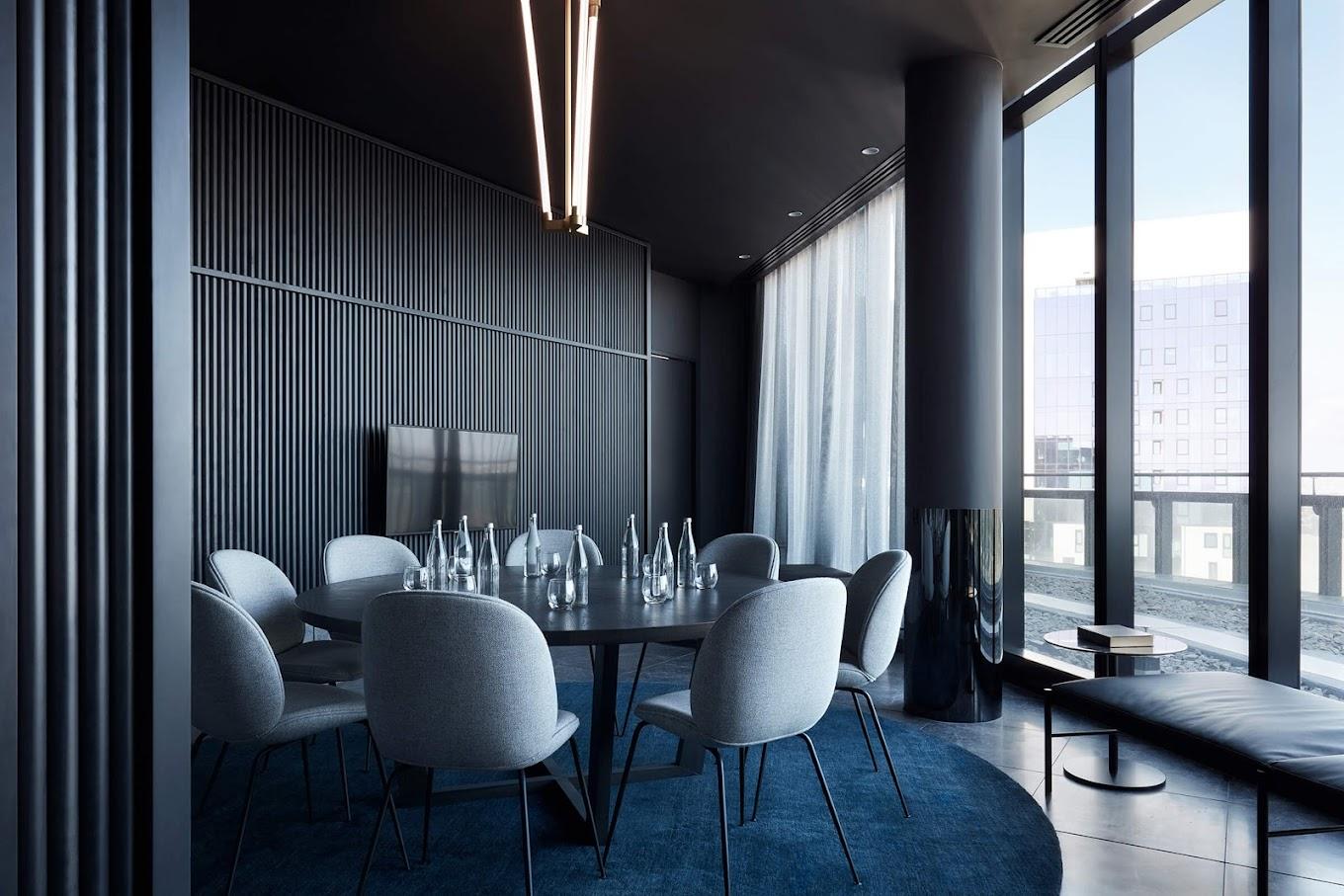 A hotel dining room with floor-to-ceiling windows, dark paneled walls, and a round table with water bottles at Shadow Play by Peppers.