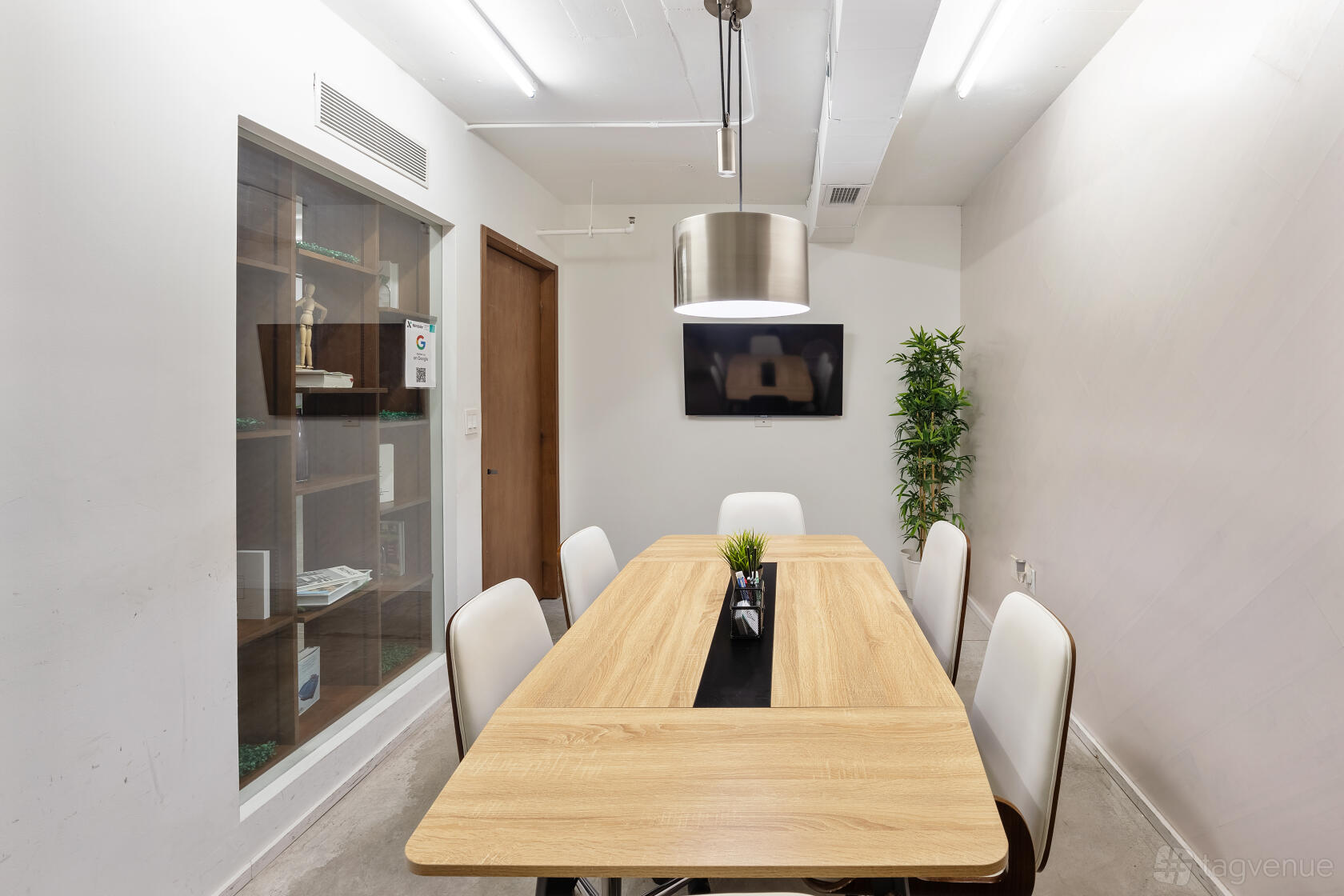 A meeting room with a wooden table, white chairs, wall-mounted TV, and a glass display cabinet at Nimbler Spaces - Miami.
