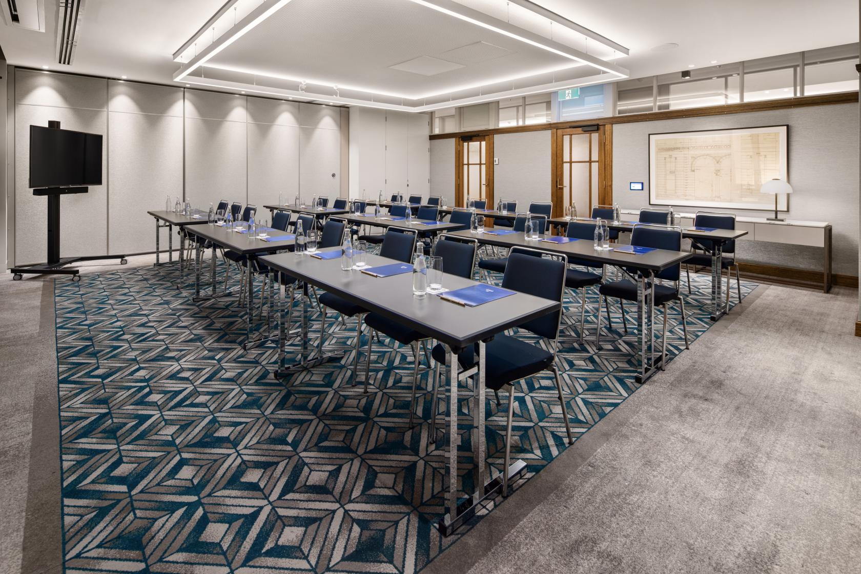 An auditorium with geometric carpet, rows of tables and chairs, and a wall-mounted screen at Hilton Melbourne Little Queen Street.