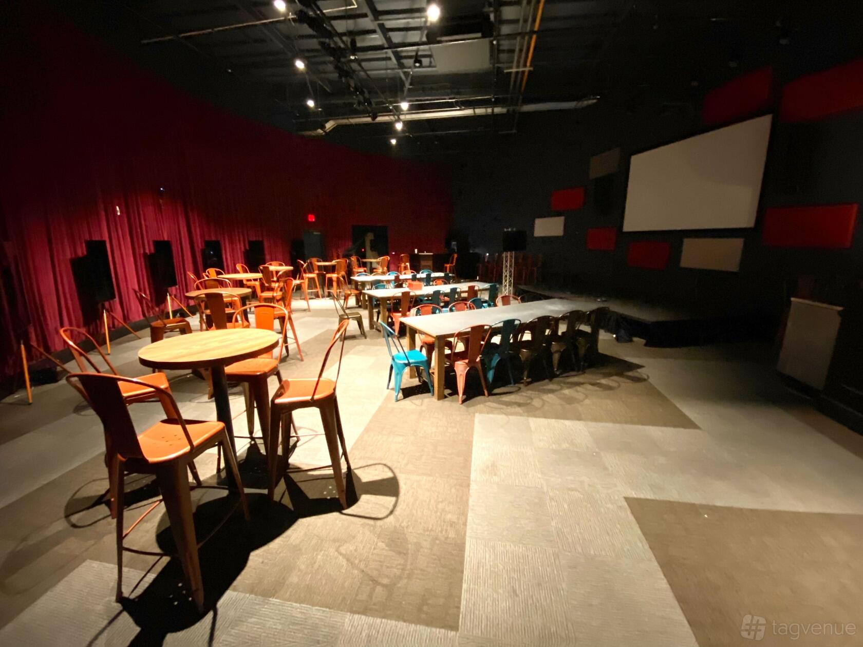 An event venue with round and rectangular wooden tables, orange chairs, and a stage with a projector screen at Two Bit Circus.
