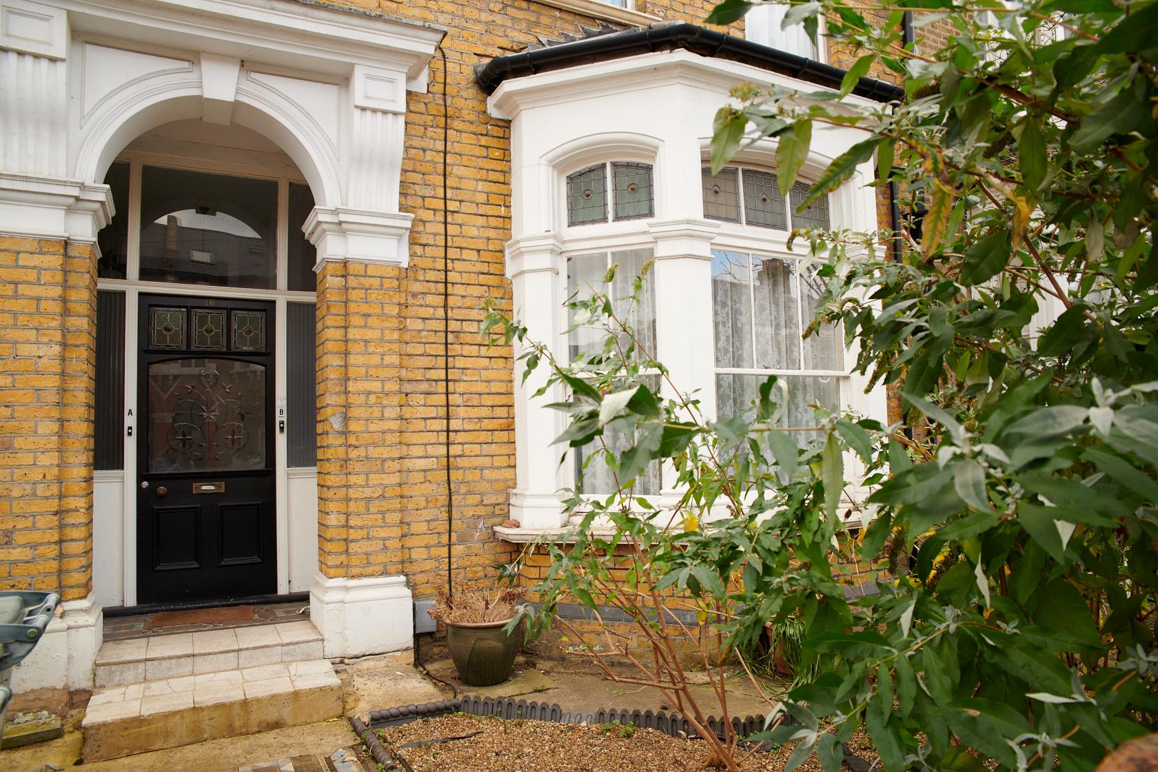 An apartment exterior with yellow brick walls, arched doorway, bay windows, and an overgrown garden at Victorian Flat with overgrown garden.