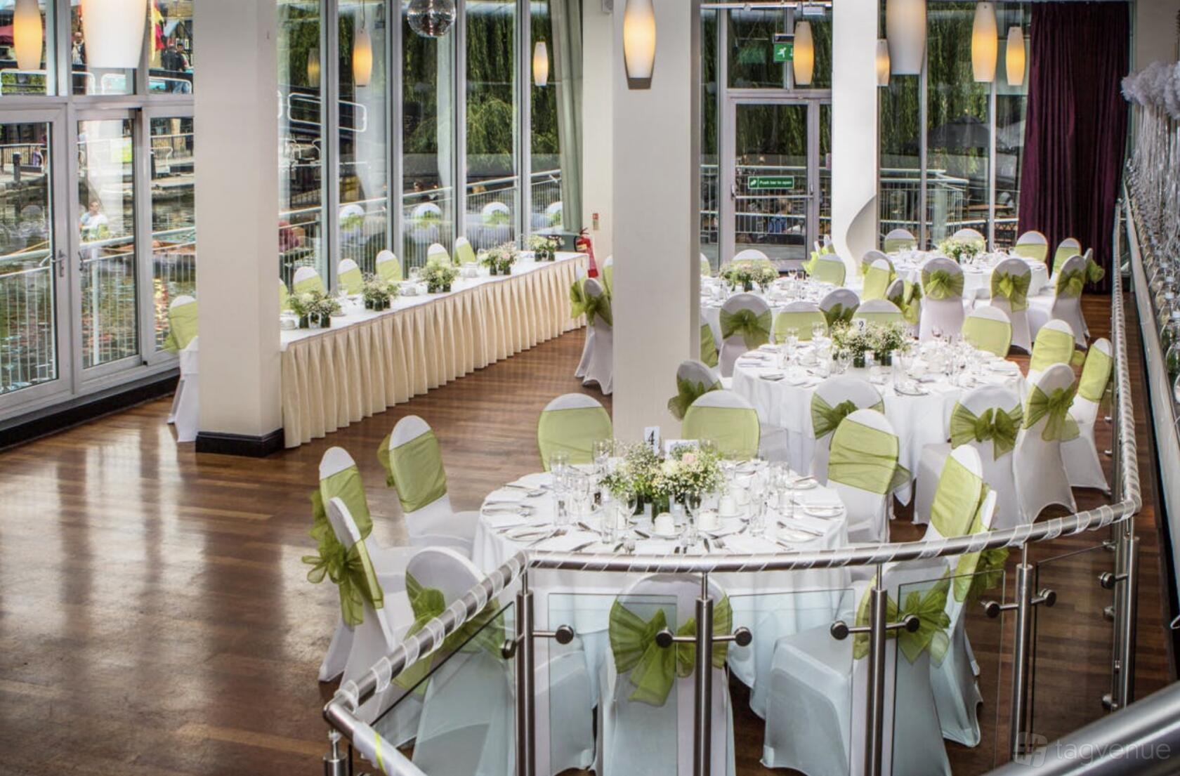 An event space with large windows, round tables with white linens and green sashes, and wooden floors at Glasshouse Camden.