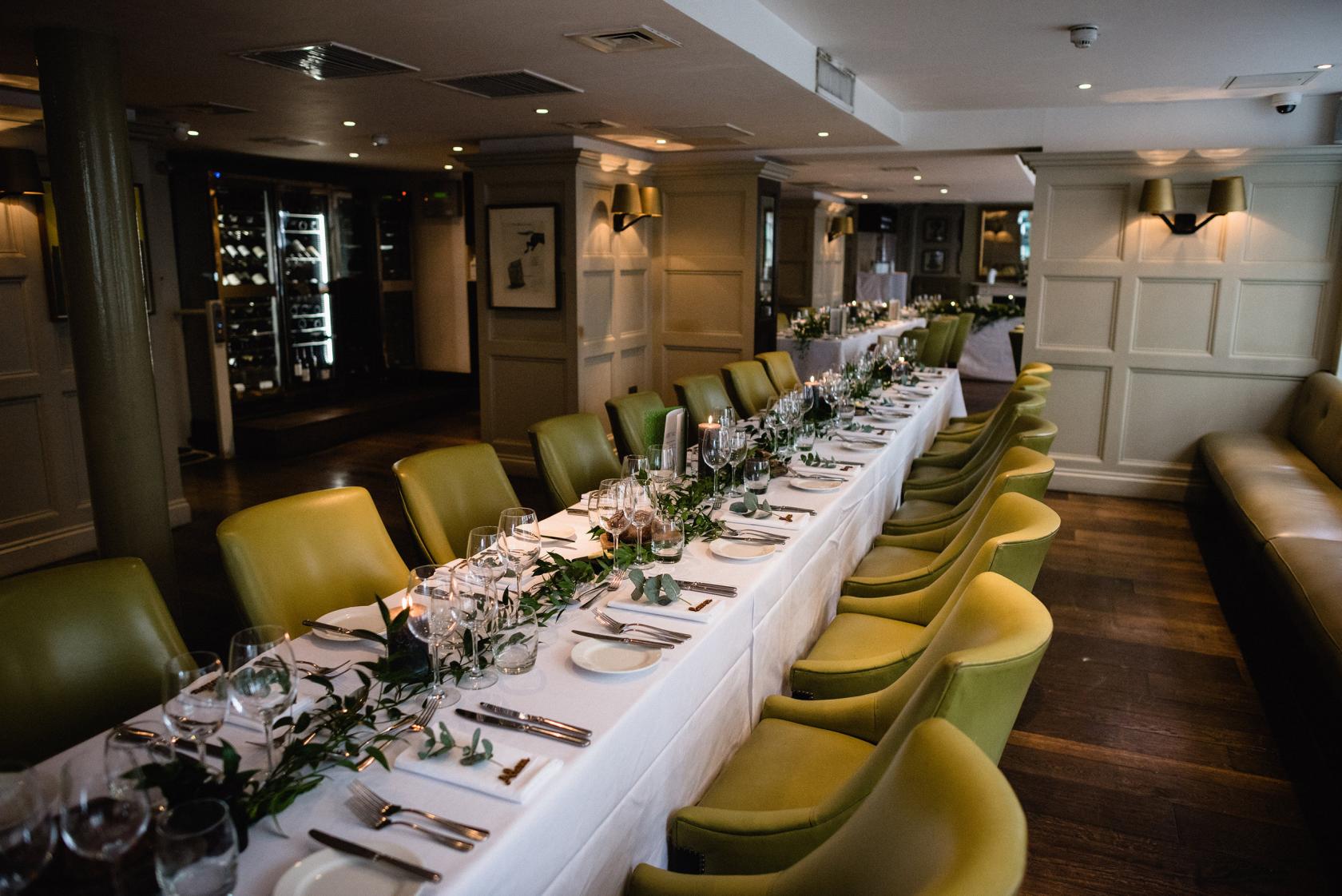 A pub with a long banquet table set with white linens, green chairs, and glassware at Chiswell Street Dining Rooms.