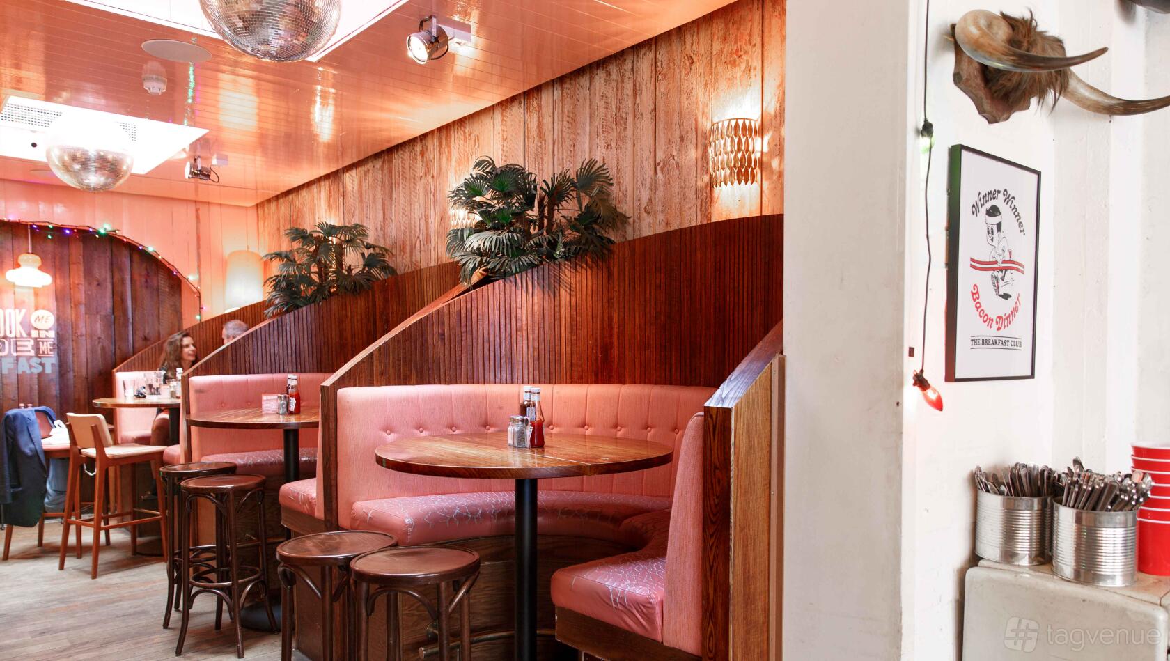 An event space in a restaurant with curved pink banquettes, round tables, and wood panel walls at The Breakfast Club Hoxton.