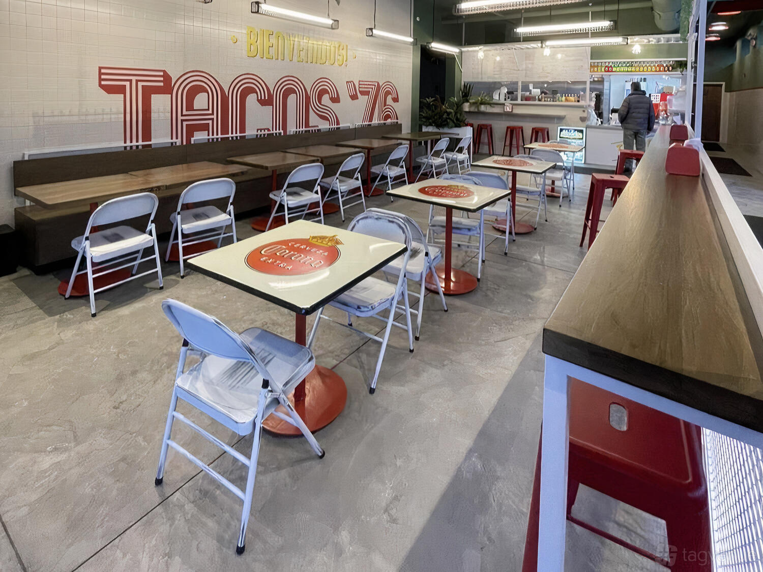 A restaurant with folding chairs, tiled floors, branded tables, and a counter at Tacos '76.