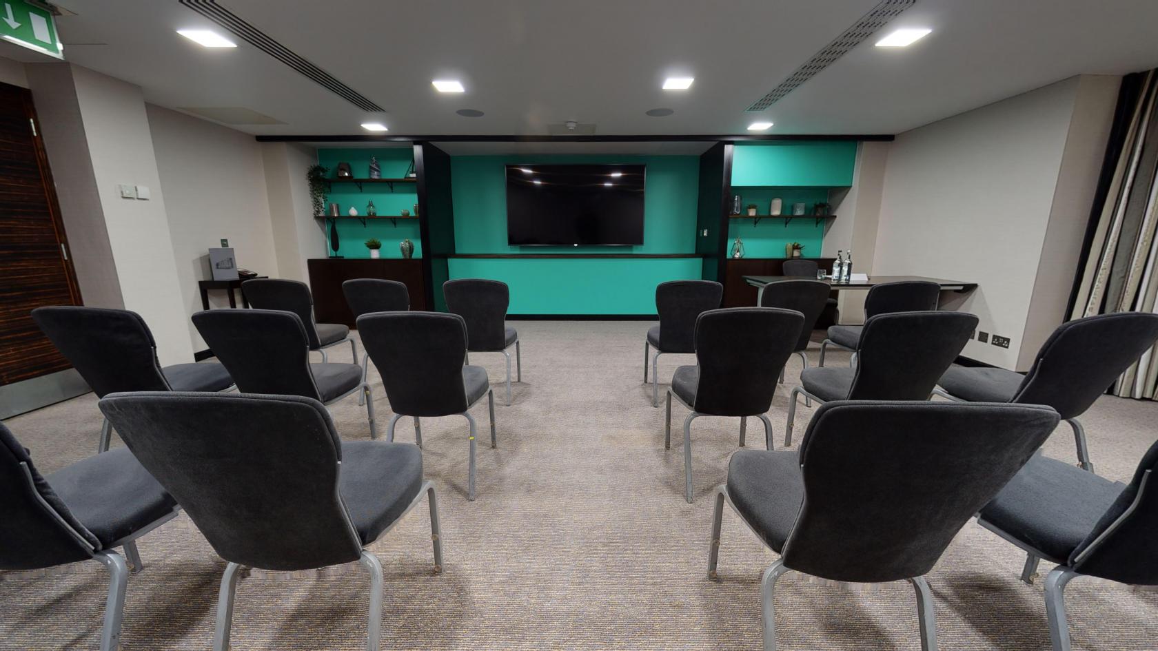 A hotel conference room with rows of black chairs, built-in shelving, and a large wall-mounted screen at DoubleTree by Hilton London Kensington.