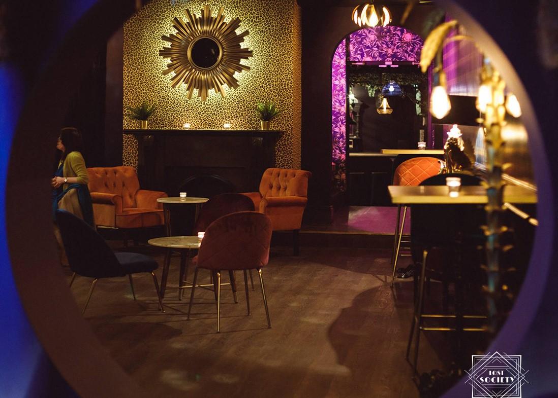 A function room with velvet chairs, round tables, and a gold sunburst mirror above a fireplace at Lost Society (Battersea SW11).