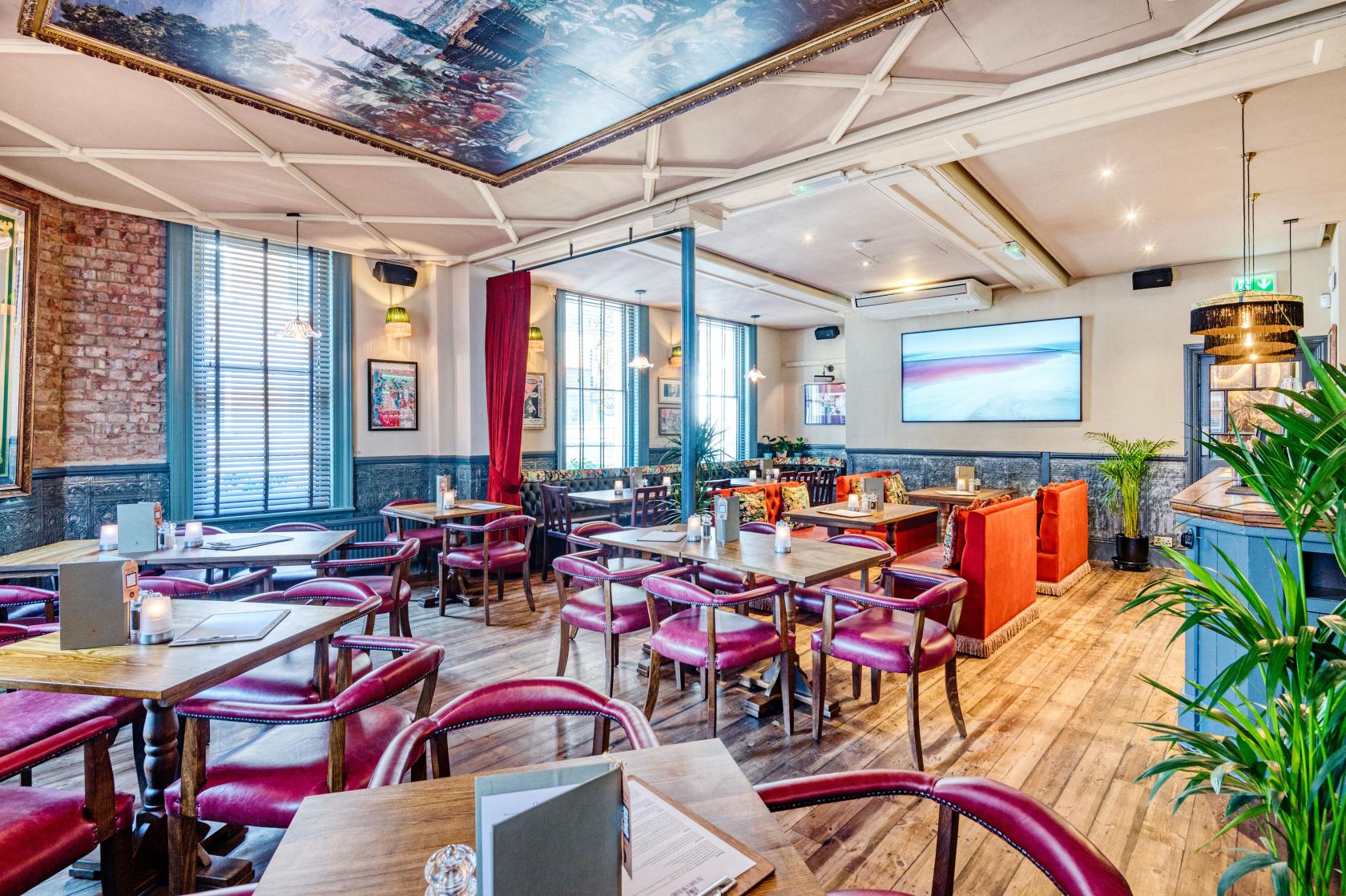 A pub lounge with wooden floors, red leather chairs, exposed brick walls, and large windows at The Crown & Two Chairmen.