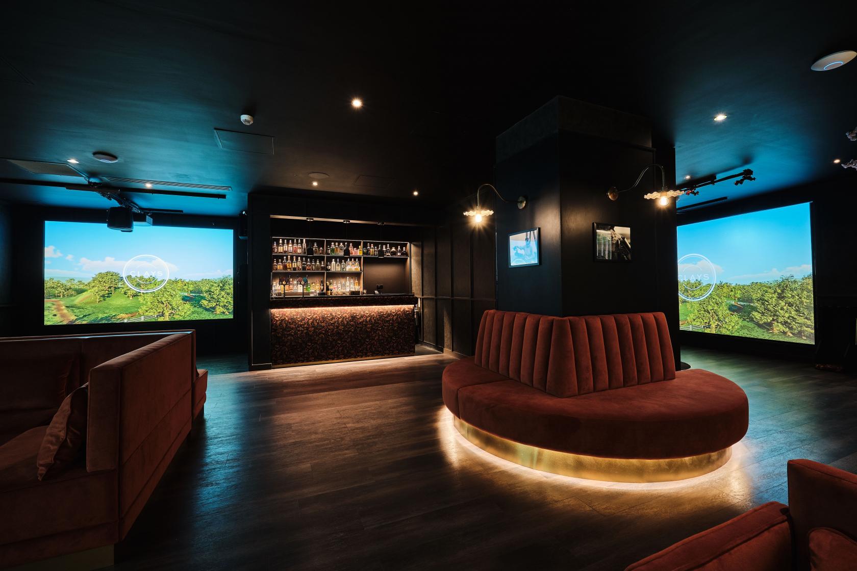 A dining room in a cocktail bar with curved velvet seating, illuminated base, and two golf simulator screens at Clays City.